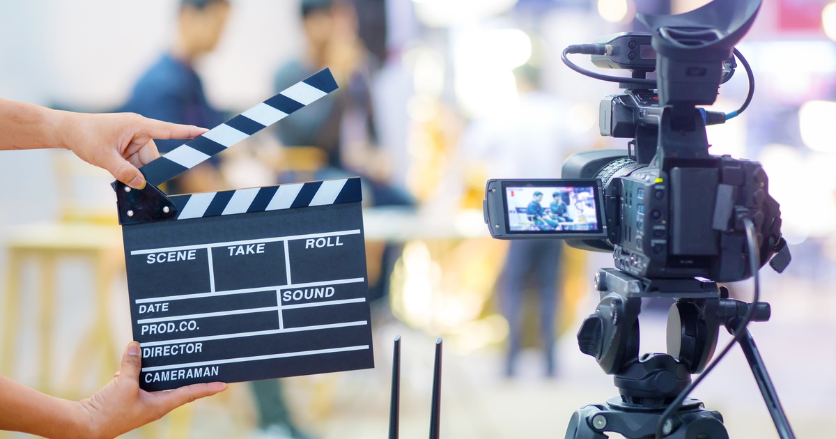 A person holding a clapperboard in front of a recording video camera as it captures two people sitting in chairs.