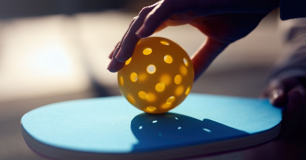 A close up of a person holding a yellow pickleball on their blue pickleball paddle. The sun is behind the paddle.