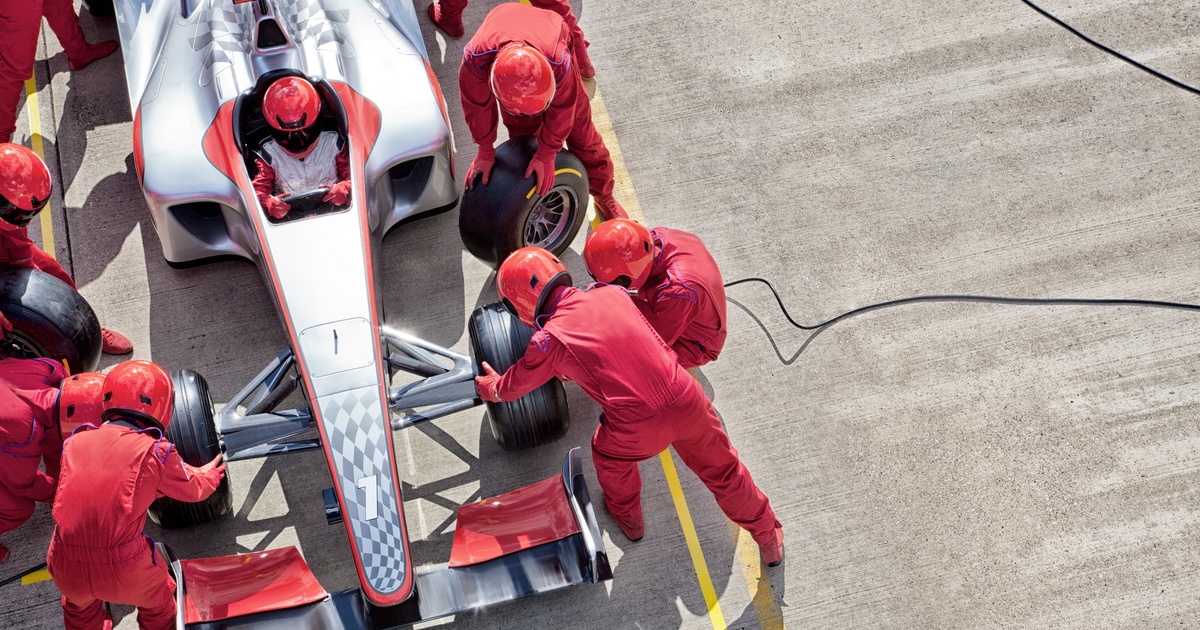 A team of eight in red coveralls changes tires on a silver Formula One car while one driver sits inside.