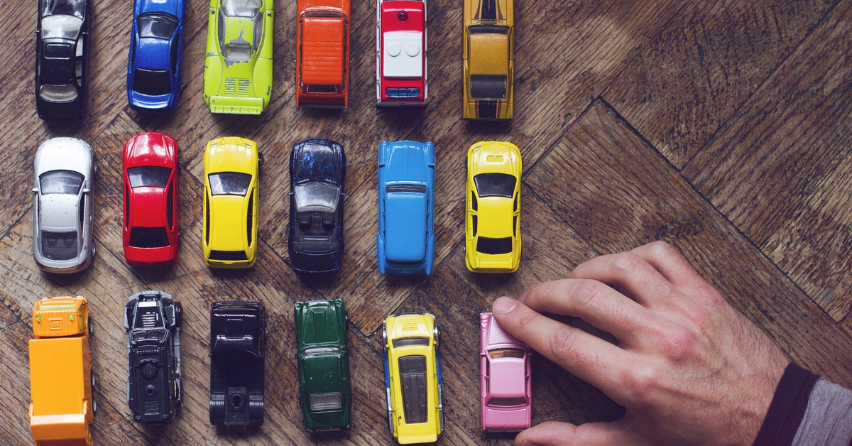 A person places a pink mini model car in their collection. The assorted colored collection sits on a wooden table.