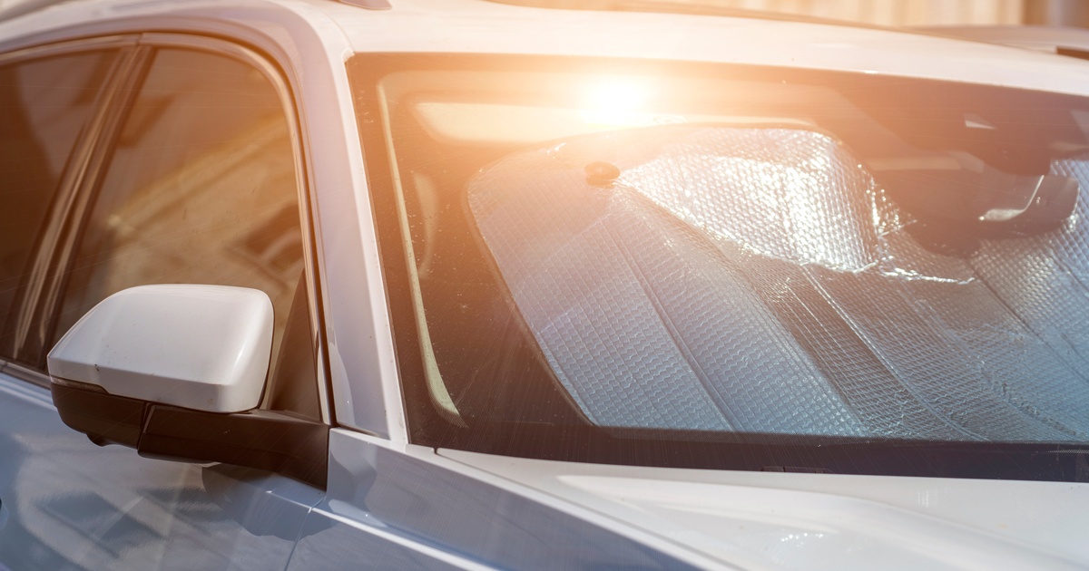 A parked car with a sun visor in the windshield sits under the hot sun. The car is silver and the windows are shut.