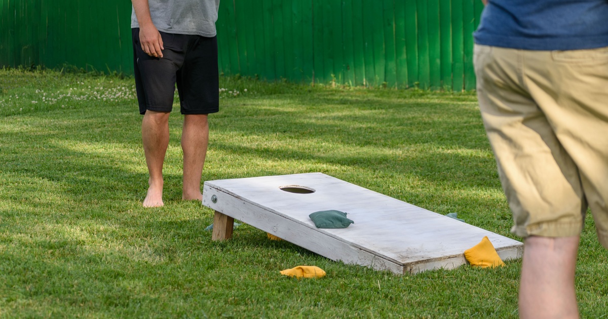 Two people stand barefoot in the grass with a cornhole board next to one of them. There are two yellow beanbags by the board.