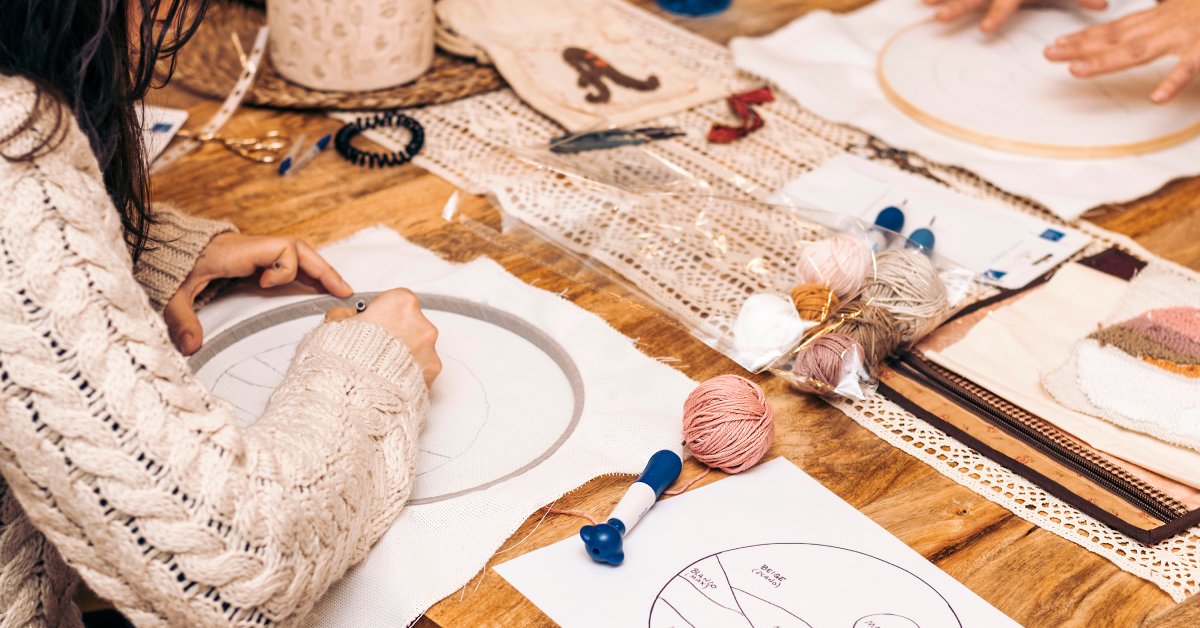A person sits at a work table with their needle stitch supplies and a project in progress. They wear a cable-knit sweater.