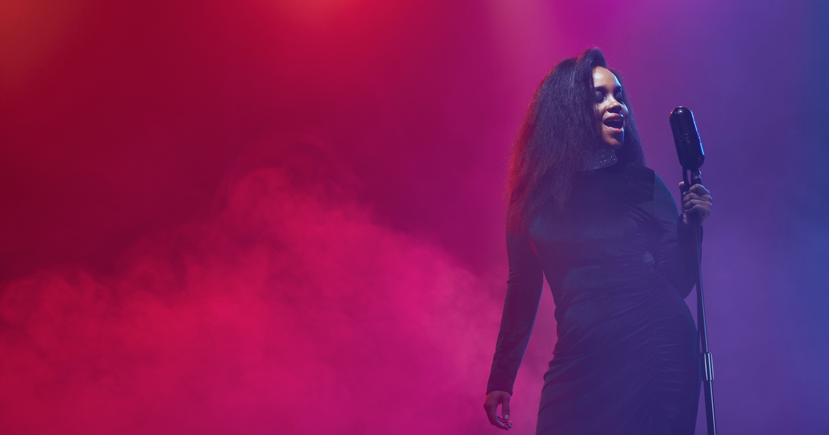 A Black woman stands by a microphone, surrounded by purple and pink lighting with fog in the background.