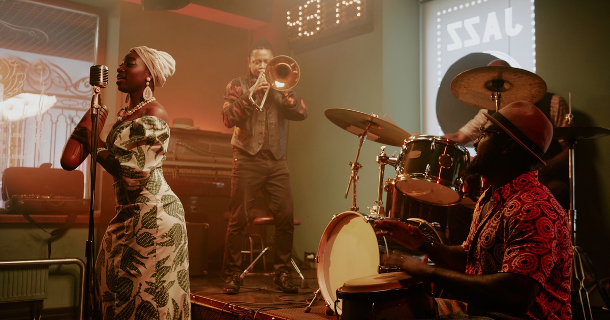 A Black woman in a green dress stands by a microphone, while two drummers and a trombone player perform on stage.