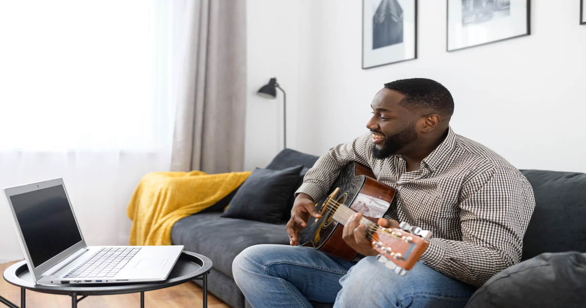 A Black man sits on his couch and plays the guitar on the video stream setup on his laptop. He's smiling as he sings.