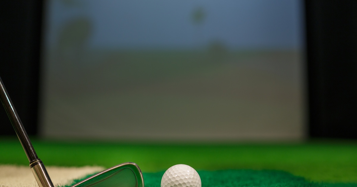 A close-up of a golf club next to a white golf ball, both resting on artificial turf, in front of a simulator screen.