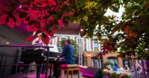 A man playing a piano on a large stage with people in the background inside a large garden next to a brick building.