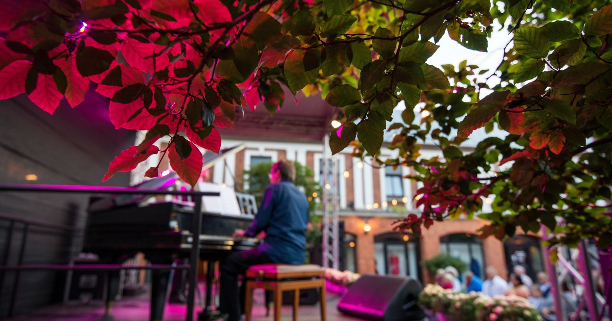 A man playing a piano on a large stage with people in the background inside a large garden next to a brick building.