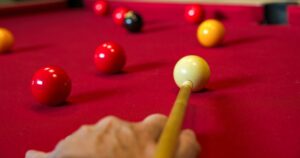 A person is aiming a pule cue at a ball on a pool table. The tables cover is red and there are several balls in play.