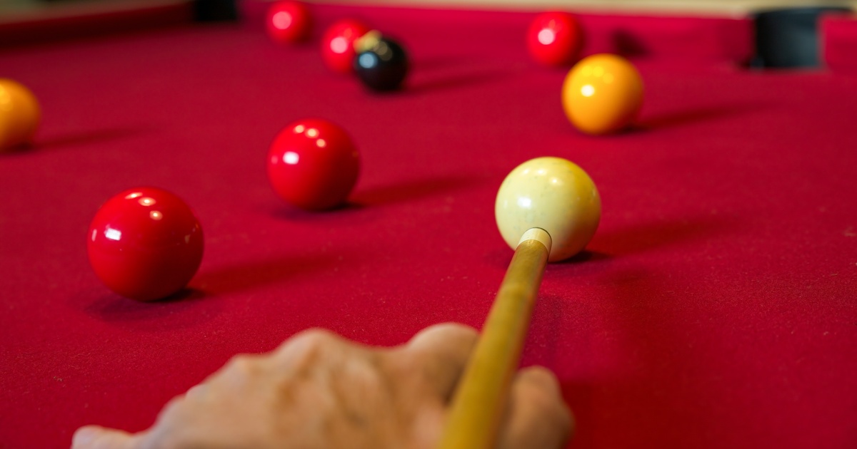 A person is aiming a pule cue at a ball on a pool table. The tables cover is red and there are several balls in play.