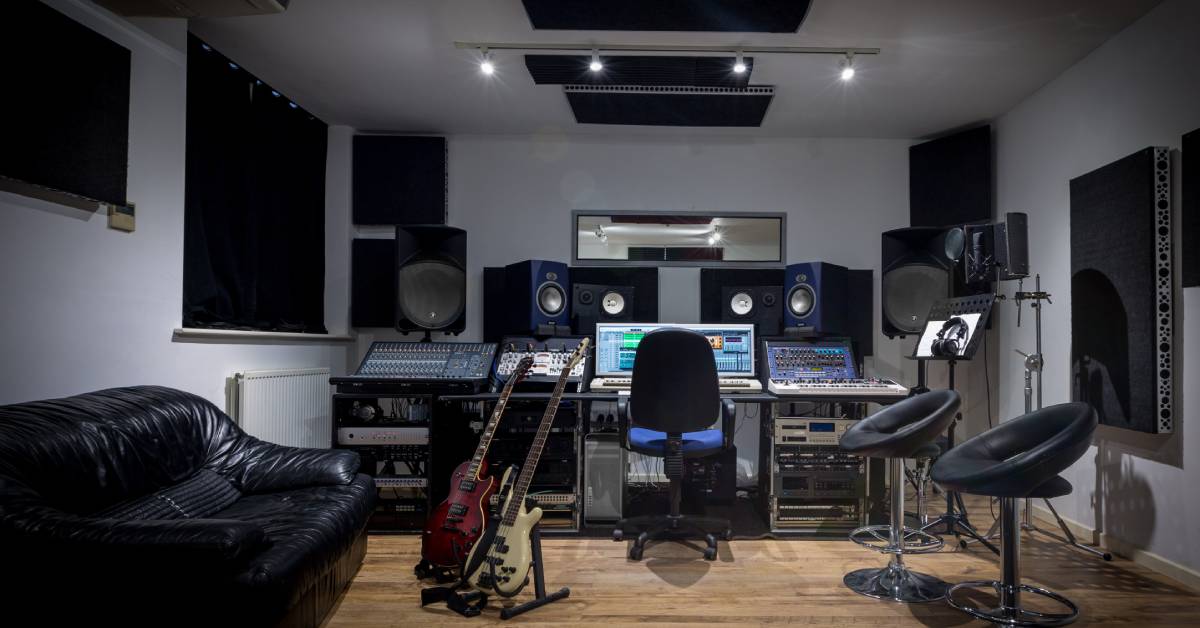 A home recording studio with two electric guitars, two stools, a couch, multiple speakers, and black curtains.