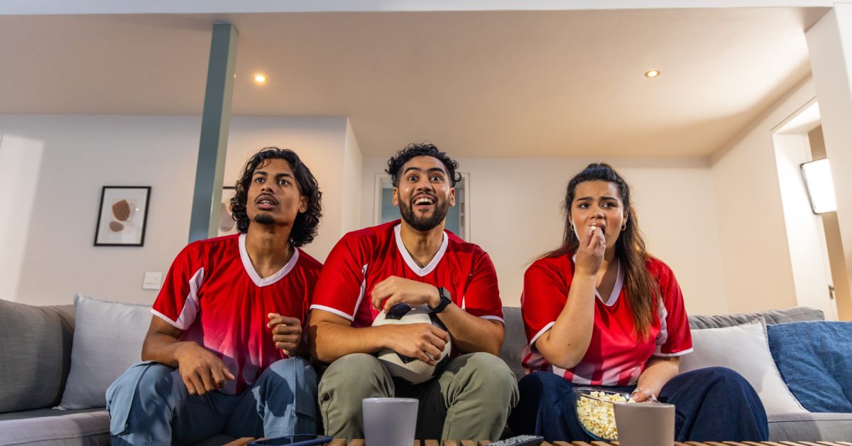 Two men and a woman wearing red sports shirts while sitting next to each other on a long couch inside a room.