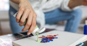 A closeup of a woman's hand squeezing green paint onto a canvas that already has red and bleu paint on it.