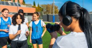 A young woman speaking into a microphone in front of female soccer players while another young woman records.