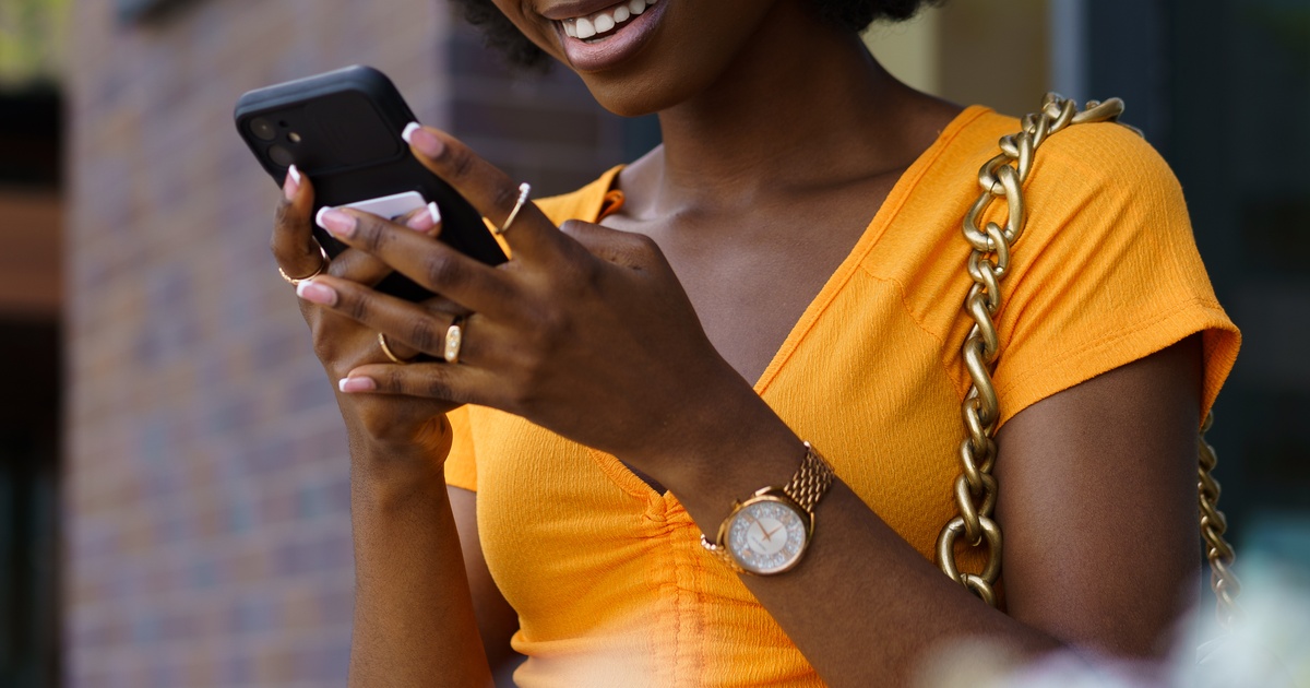 A woman in an orange top smiles as she uses her phone. She wears a gold watch, rings, and a chunky chain-strap bag.