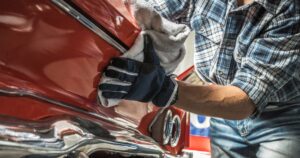A man in a blue plaid shirt is using a towel to clean the rear fender of a car. The car is red and glossy.