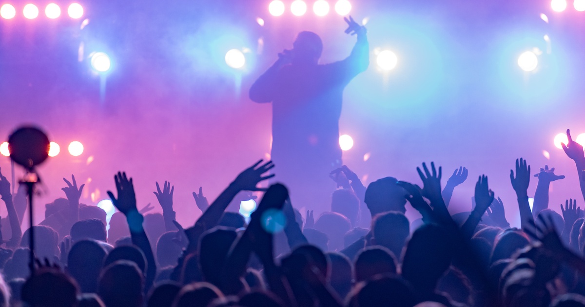 A rapper performs on stage under bright lights as the crowd raises their hands in a hazy concert venue.