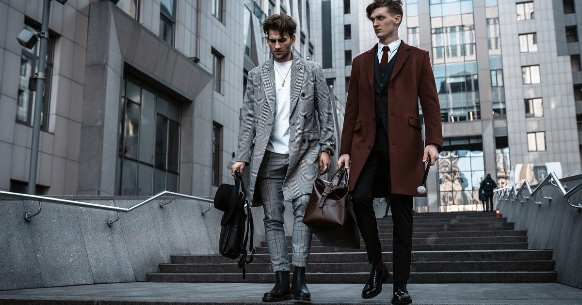 Two men wearing suits and coats with black shoes while carrying large leather bags walking down street steps.