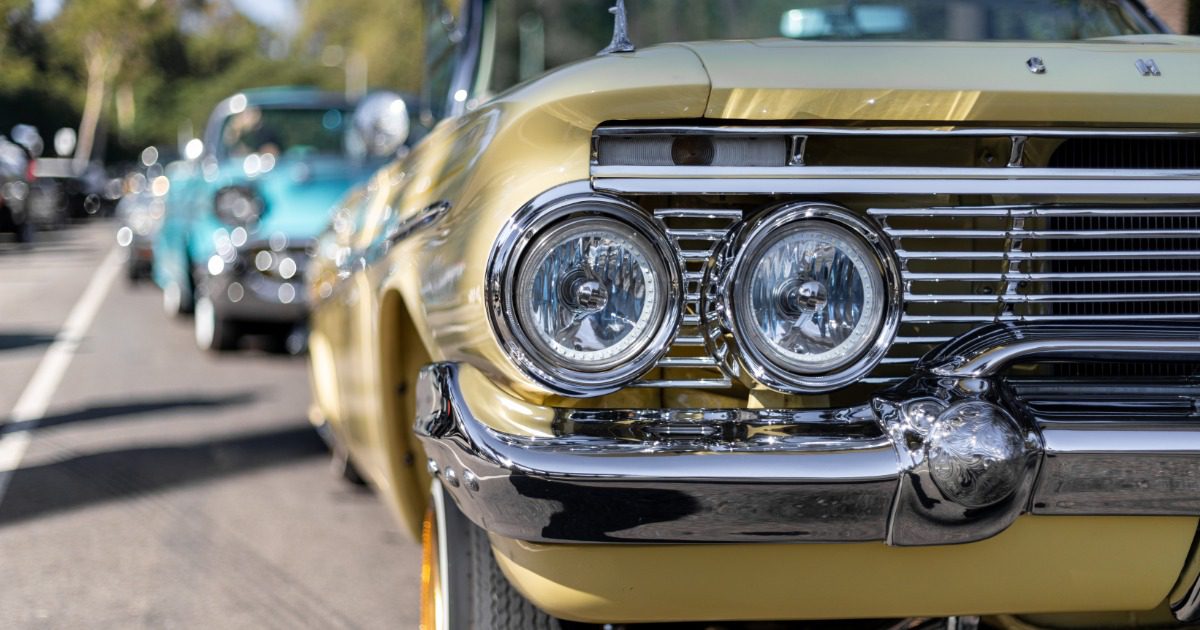 A yellow lowrider with a chrome grille and round headlights parks on a sunny street with classic cars parked behind it.