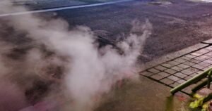 A manhole cover releases steam on a dark city street beside a paved sidewalk and a green metal barrier.