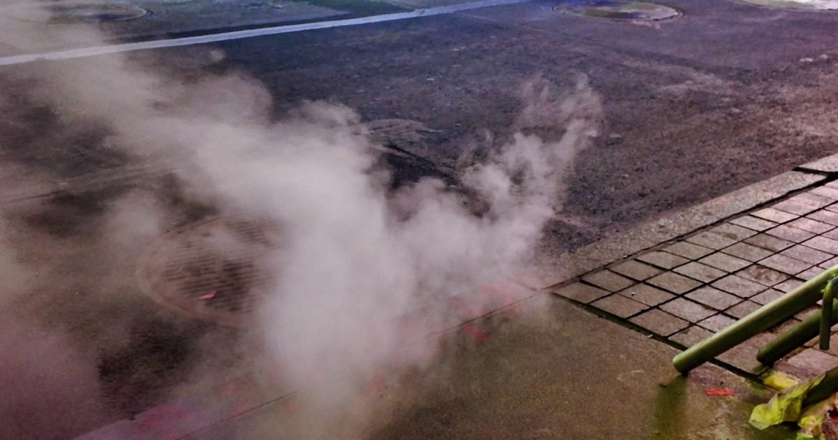 A manhole cover releases steam on a dark city street beside a paved sidewalk and a green metal barrier.