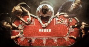 Five players gather around a red poker table as a dealer holds cards among colorful chips, drinks, and smoky lights.