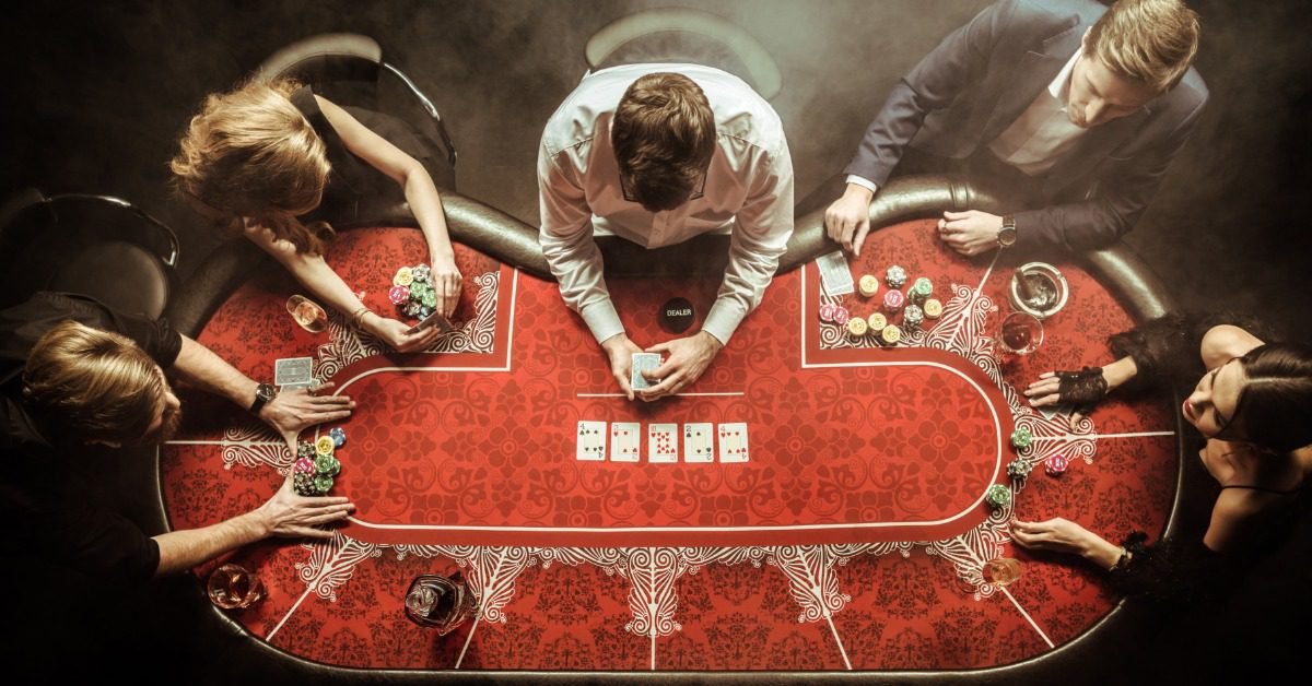 Five players gather around a red poker table as a dealer holds cards among colorful chips, drinks, and smoky lights.