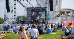 People are sitting on a grassy lawn by a stage. Speakers, lights, musical instruments, and a screen are on the stage.
