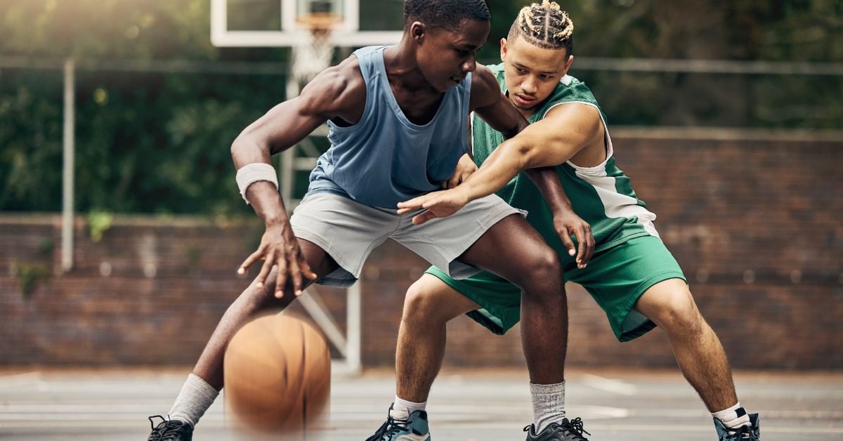 Two basketball players fight for control of the ball on an outdoor court, with trees and a brick wall in the background.